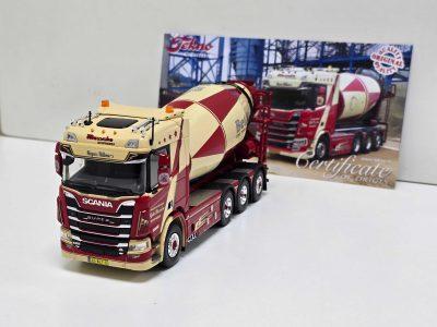Scale model of a red and beige Scania cement mixer truck on a white surface with a background certificate poster behind it.