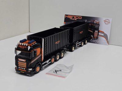 Scale model Scania toy truck with orange and black trailers on a white surface, with a small bag of screws in the foreground and a Henco Trans backdrop in the background