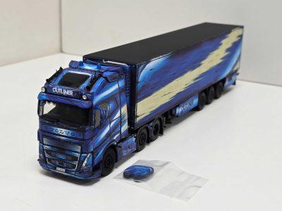 Blue toy Volvo truck with trailer on a white surface, a round blue token nearby resting on a folded envelope or paper folder.
