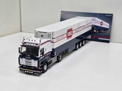 Scale-model Scania truck with a long white trailer labeled Van Dijk Holland and a blue cab, displayed on a white surface against a backdrop poster in the background.