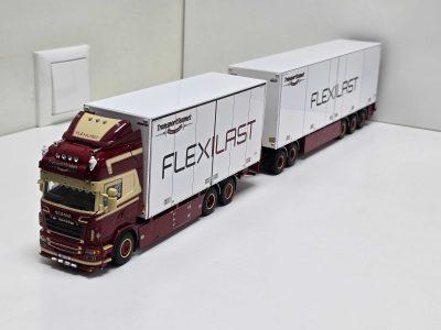 Red toy truck pulling two white trailers labeled FLEXILAST indoors on a white surface.