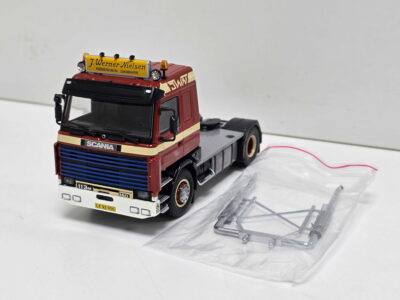 Red scale-model truck cab with a yellow sign on top and loose plastic trailer parts nearby on a white surface