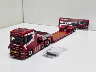 Red toy truck with attached trailer on a white surface, front labeled Scania/J26 Trucking, with a red trailer and a small instruction/packaging card in the background.