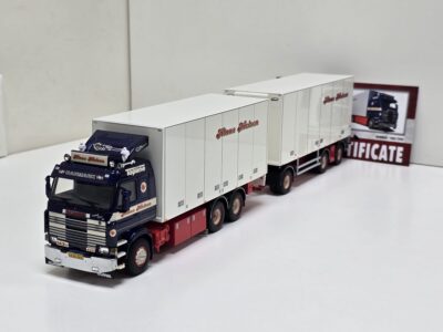 Scale model trucks lined up on a white surface, blue Scania cab in front and white trailers labeled 'Haus Nielsen' behind it