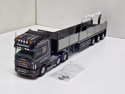 Scale model of a black Scania truck with a long gray trailer and crane attachments on a white surface, with plastic bags nearby.”] ,