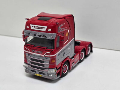 Red Scania toy truck model with a gray bumper and decorative decals, parked on a white surface on a slight angle.