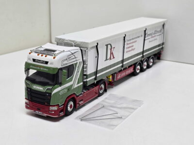 Scale-model Scania truck with a long white trailer in a white studio setting, showing detailed cab paintwork in green and red and brand logos on the side men.