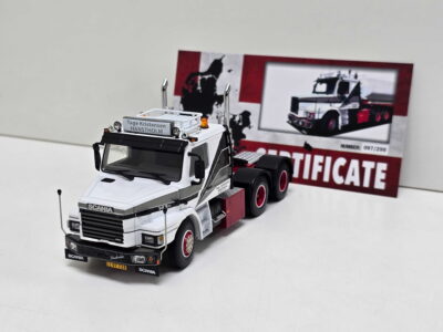 Close-up of a white Scania toy truck with a yellow license plate on a white surface, foregrounding the front grille and signage reading Tage Kristensen HANSTHOLM.
