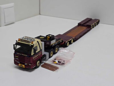 Model truck with a long brown trailer loaded with multiple flatbed sections, on a white surface next to plastic bags of small brown items, view from front-left corner to right.