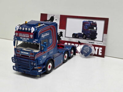 Blue toy Scania truck model with front bumper guard and multiple roof lights, placed in front of its packaging box.
