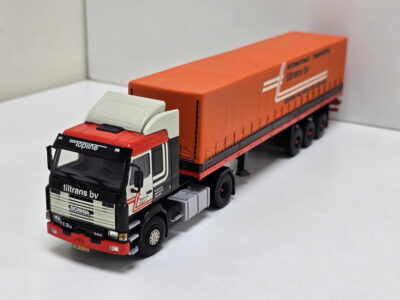 Scale-model Scania truck with a long orange trailer on a white surface, close-up of the cab in foreground and detailed branding on the trailer edge.