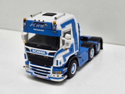 Close-up of a blue-and-white Scania toy truck cab, showing logo, headlights, and orange indicator lights against a plain background.