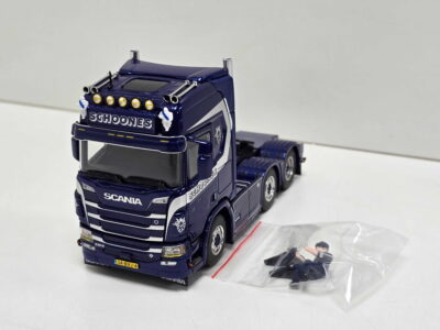 Blue toy Scania truck model with multiple chrome headlights and 'SCHOONES' branding on the front, placed on a white surface beside a clear plastic bag with figures.