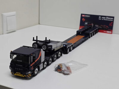 Black Scania toy truck with a long flatbed trailer in a white room; a bag of small parts sits on the floor nearby and a sign is in the background.
